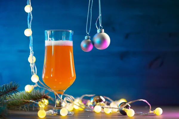 A glass of festive Christmas beer ale on a wooden blue background. Christmas tree branches and a round yellow garland. Copy space