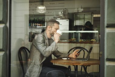 Kafkasyalı yüzü olmayan bir adam bir kafede camın arkasında dizüstü bilgisayarla çalışıyor. Adam kahve içiyor ve telefona bakıyor. Çoklu tarama kavramı