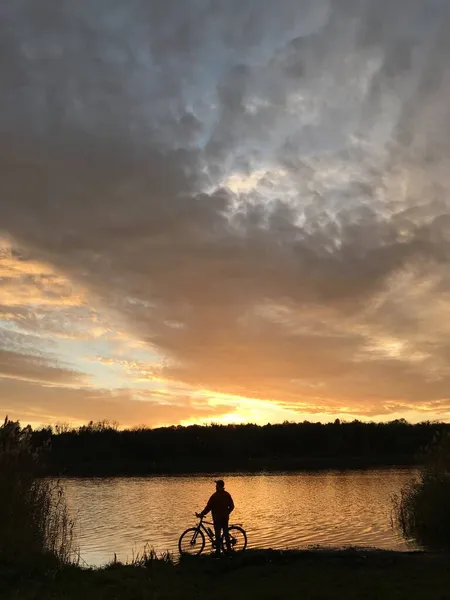 Silueta de un hombre con una bicicleta en el fondo del lago y puesta de sol. — Foto de Stock