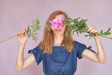 Mavi elbiseli yüzü olmayan kızıl saçlı bir kadın yüzünü açık bir duvara karşı pembe şakayıklıklarla kaplıyor. bahar konsepti 