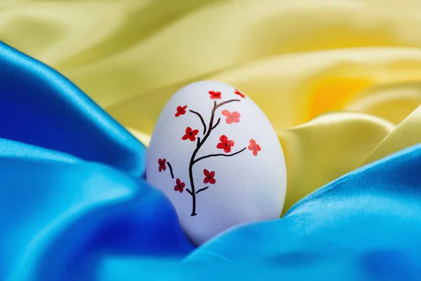 Beyaz Paskalya yumurtası, Ukrayna bayrağının mavi ve sarı saten arka planında güzel boyanmış kırmızı çiçekler. Savaş sırasında bir tatil..
