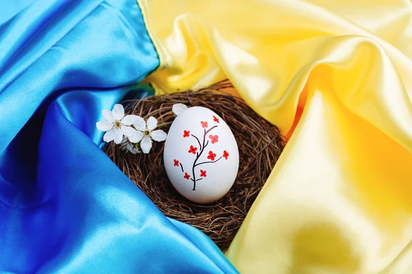 白色的复活节彩蛋 色彩艳丽的红花插在乌克兰国旗蓝色和黄色明亮缎子背景上的木制柳条鸟巢中 在占领期间对该国的支持 — 图库照片