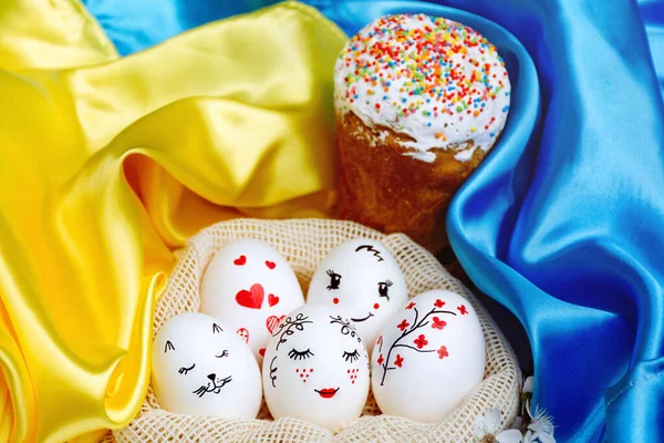 White Easter eggs in a bag with beautiful painted faces and a delicious Easter cake with colorful sweet fillings and a glaze of blue and yellow bright satin background of the State Flag of Ukraine..