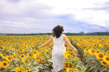 Beyaz elbiseli, kıvırcık saçlı güzel kız sarı ayçiçeği tarlasında koşuyor. Fırtınadan önce mavi kasvetli gökyüzü. Fotoğraf üzerine yumuşak seçici odak.