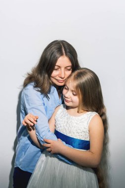 Mavi gömlekli anne, güzel, uzun sarı saçlı ve açık arkaplanda mavi kurdeleli zarif beyaz elbiseli kızına sarılıyor. Doğum günü ya da Anneler Günü.