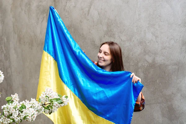 Gülümseyen kız, Ukrayna 'nın mavi ve sarı bayrağını beton bir duvarın arkasında tutuyor. Kareli gömlekli bir kadın. Ağaç dalları filizlenir. Uzayı kopyala Anayasa Günü.