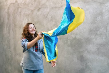 Ukrayna 'nın mavi ve sarı ulusal bayrağını tutan gülümseyen kıza odaklanın. Arka planda desenli beton duvarda geleneksel işlemeli gömlekli bir kadın. Bağımsızlık Günü. Boşluğu kopyala