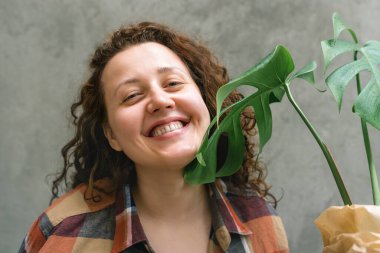 Mutlu, samimi, gülümseyen kadın dik duruyor ve elinde yeşil bir bitki tutuyor. Beton duvarda kareli gömlekli kıvırcık saçlı bir kız. Doğal güzellik kavramı.