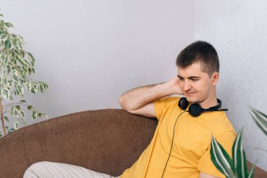 Evde sarı tişörtlü, kulaklık takan genç adam. Çocuk müzik ya da konferans dinler. Odada yeşil bitkiler var. Çevrimiçi eğitim ve ev bahçesi kavramı.