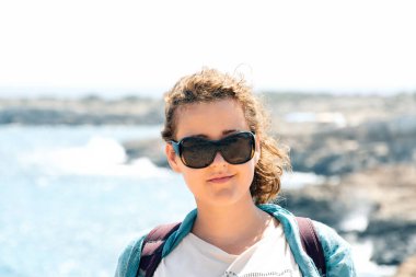 Genç, gülümseyen bir kadın, sıcak ve güneşli bir günde mavi denizin arka planına ve kayalara bakıyor. Güneş gözlüklü kadın, sırt çantası. Turist bayramının açılışı. Uzayı kopyala İyi istirahatler.