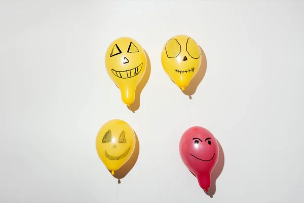 Four scary balloons painted for Halloween hanging on white wall background. One stands out red among yellow-orange ones. Different emotions of joy, anger, laughter. Holiday decoration, party concept.