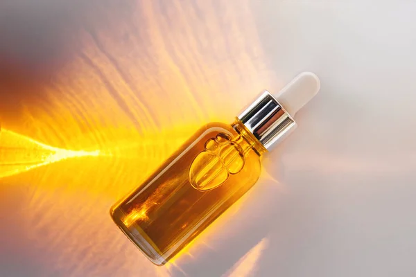 Mockup of a glass dropper bottle with a metal cap and yellow essential oil on a background with an abstract fire pattern of light and shadow. Eco cream, serum, cosmetics, massage oil. Copy space