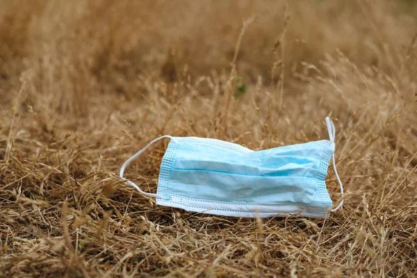 Waste during COVID-19. Blue Medical face mask thrown into nature in yellow grass. End of quarantine. Environmental pollution with plastic.