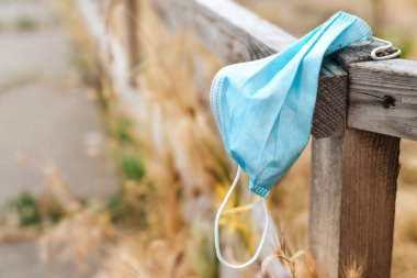 Waste during COVID-19. A blue medical face mask hangs on a wooden fence and is thrown into the nature among the yellow grass. End of quarantine. Environmental pollution with plastic.