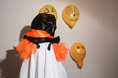 Mutlu Cadılar Bayramı. Ev yapımı çarşaftan yapılmış ev yapımı bir kostümle duvarda turuncu toplarla boyanmış beyaz bir hayalet. Şenlik tasarımı, parti konsepti. Azizler Günü kutlaması.