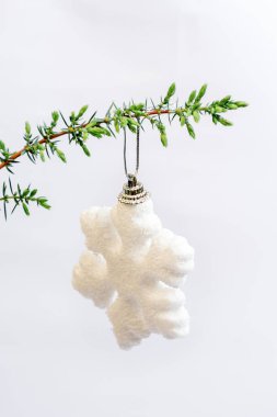 Soft selective focus on white snowflake Christmas toy hanging on green branch of Christmas tree, juniper on white background. Minimal style. Happy holidays. Vertical Photo. Copy space