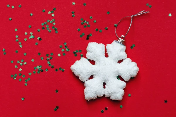 One White big snowflake and small green twinkling decorative shiny stars shine on red surface. View from above. Christmas and new year celebration concept. Copy space for text