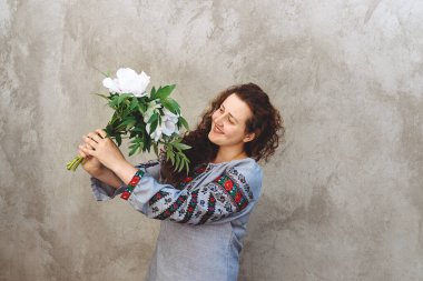Girl with bouquet of white peonies. Woman wears traditional embroidered Ukrainian shirt against concrete wall. Woman is smiling. Independence Day of Ukraine. Copy space. Soft focus