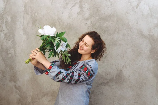 Girl with bouquet of white peonies. Woman wears traditional embroidered Ukrainian shirt against concrete wall. Woman is smiling. Independence Day of Ukraine. Copy space. Soft focus