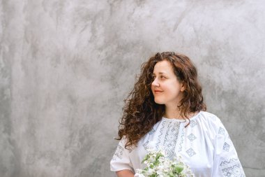 Smiling woman with long curly hair in traditional white embroidered Ukrainian shirt against concrete wall background. The girl looks away. Independence Day of Ukraine. Copy space. Soft focus