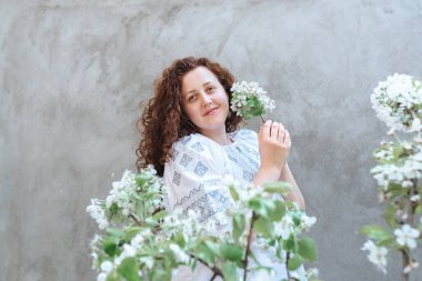 Woman long curly hair in white embroidered Ukrainian shirt. Defocused focus on foreground on blossoming branches of apple tree on background of concrete wall. Independence Day of Ukraine. Soft focus