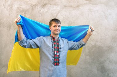 Young man holds blue-yellow flag of Ukraine above his head. Man in traditional embroidered shirt against background of textured concrete wall. Front view, copy space. Independence Day of Ukraine