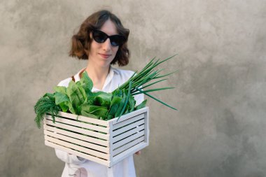 Defocused focus on girl holding a white wooden box full of fresh vegetables - spinach, onions, dill on concrete wall background. Woman in sunglasses. Ideal for dietary spring salad. Copy space