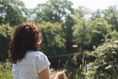 Girl traveler sits on grass among green trees and holds blade of grass. Woman with curly hair. Personal pleasure of solitary silence. Digital detox. Copy space. Soft selective focus