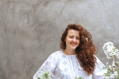 Woman with long curly hair in white embroidered shirt. Defocused focus in foreground on blossoming branches of apple tree against background of concrete wall. Independence Day of Ukraine. Soft focus