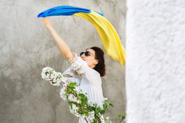 Soft Selective focus on Girl waving blue and yellow Flag of Ukraine. Woman in white dress and sunglasses in city. Defocused focus on foreground Apple and pear blossoms. Copy space. Independence Day