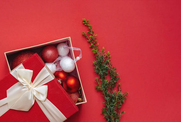 Christmas decorations in open gift box and green Christmas tree branch or juniper on plain background. Red packaging lid with lush bow. Merry Christmas and Happy New Year. Copy space.