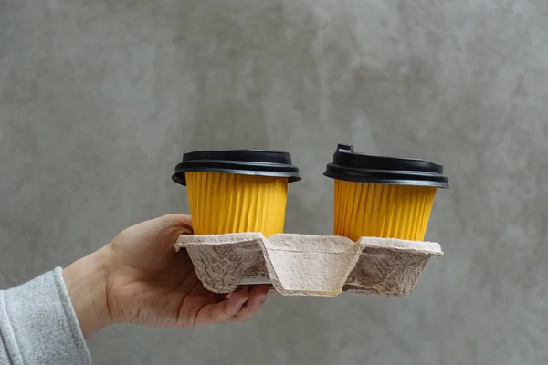 Two yellow cups with hot takeaway coffee in paper holder in female hand on background of concrete wall in city. Morning pleasure before work. Copy space for text