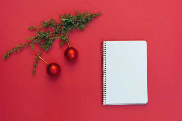 Kırmızı arka planda süslemeleri olan Noel ağacı dalının yakınındaki Açık Boş Spiral defter üzerine yumuşak seçici bir odaklanma. Uzay metnini, yapılacaklar listesini, rüyaları kopyala. Planlama 2023, Noel dilekleri listesi