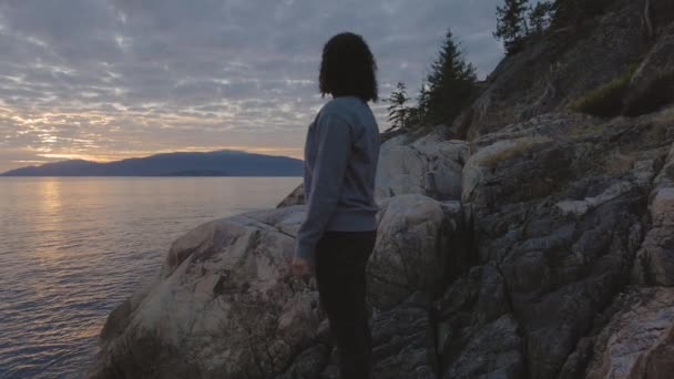 Vit kvinna på en stenig kust under en dramatisk grumlig solnedgång. — Stockvideo