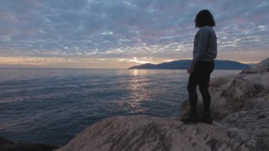 Kayalık sahildeki beyaz kadın dramatik bulutlu bir günbatımında.