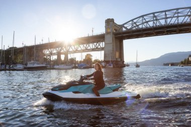 Okyanusta Sulu Scooter süren Maceracı Kafkas Kadın. Güneşli Günbatımı Gökyüzü. Vancouver şehir merkezi, British Columbia, Kanada. Burrard Köprüsü.