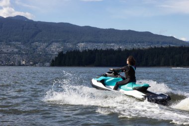 Adventurous Caucasian Woman on Water Scooter riding in the Ocean. Modern City and mountains in background. Downtown Vancouver, British Columbia, Canada.