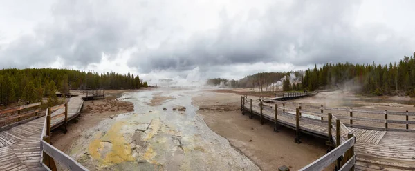 Wooden hiking path in Hot spring Geyser with colorful water in American Landscape. Cloudy Sky. Yellowstone National Park, Wyoming, United States. Nature Background Panorama