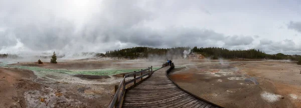 Wooden hiking path in Hot spring Geyser with colorful water in American Landscape. Cloudy Sky. Yellowstone National Park, Wyoming, United States. Nature Background Panorama