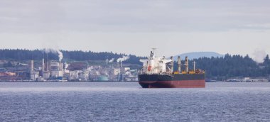 Industrial Processing Plant and Container Ship passing by on a cloudy day. Nanaimo, Vancouver Island, British Columbia, Canada.