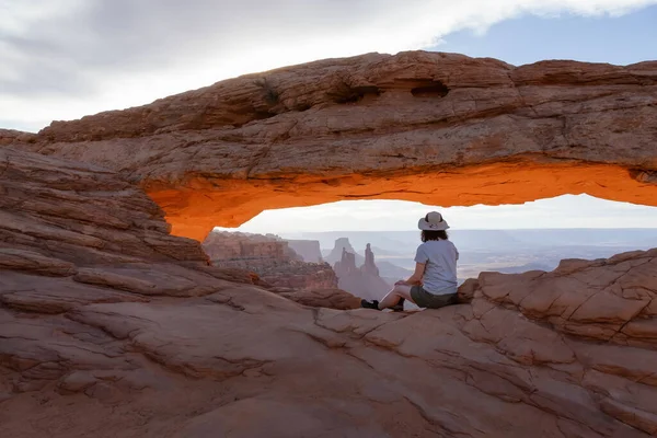 Adventurous Woman at a Scenic American Landscape and Red Rock Mountains in Desert Canyon. Spring Season. Sunrise Sky. Mesa Arch in Canyonlands National Park. Utah, United States. Adventure Travel