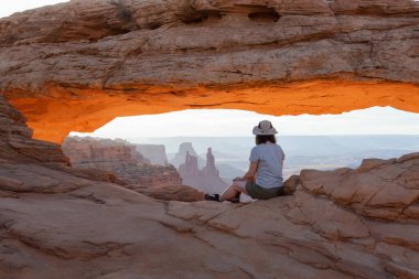 Adventurous Woman at a Scenic American Landscape and Red Rock Mountains in Desert Canyon. Spring Season. Sunrise Sky. Mesa Arch in Canyonlands National Park. Utah, United States. Adventure Travel