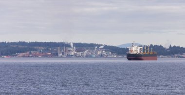 Industrial Processing Plant and Container Ship passing by on a cloudy day. Nanaimo, Vancouver Island, British Columbia, Canada.