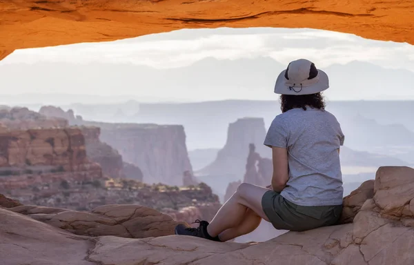 Adventurous Woman at a Scenic American Landscape and Red Rock Mountains in Desert Canyon. Spring Season. Sunrise Sky. Mesa Arch in Canyonlands National Park. Utah, United States. Adventure Travel