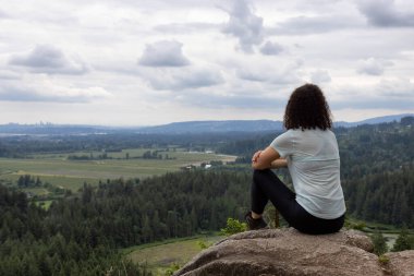 Adventurous Woman Standing on top of a rock overlooking the Canadian Nature Landscape. Minnekhada Regional Park, Coquitlam, Vancouver, British Columbia, Canada.