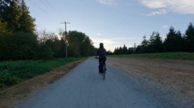 Yeşil ağaçlarla çevrili bisiklet yolunda bisiklet süren maceraperest kadın. Yaz Günbatımı. Pitt Meadows, Greater Vancouver, British Columbia, Kanada.