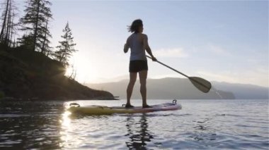 Huzurlu bir gölde kürek çeken maceraperest bir kadın. Güneşli Günbatımı. Harrison Kaplıcaları, British Columbia, Kanada. Macera Sporu Seyahat Konsepti. Yavaş Sinema Sineması