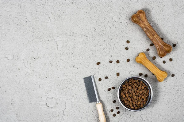 Dry dog pet food in bowl and accessories on gray concrete background top view. Pet feeding and care concept background with copy space. Photograph taken from above.