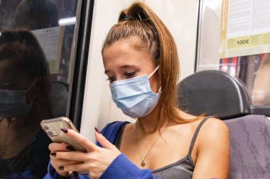 Barselona, İspanya - 21 Eylül 2021: Genç bir kadın metroda seyahat ederken cep telefonunu kullanıyor. Koronavirüs covid-19 salgınından dolayı koruyucu yüz maskesi kullanıyor.