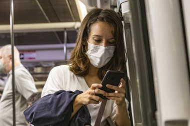 Barselona, İspanya - 21 Eylül 2021: Bir kadın metroda seyahat ederken cep telefonunu kullanıyor. Koronavirüs covid-19 salgınından dolayı koruyucu yüz maskesi kullanıyor.